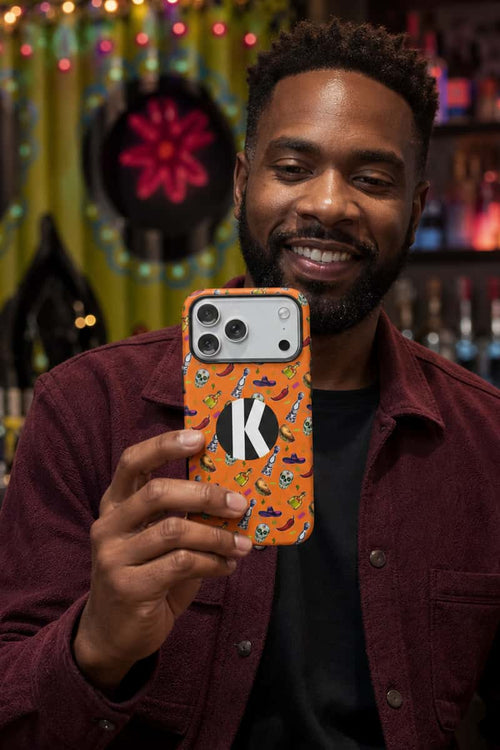 A man holding up an orange Halloween-themed phone case with a black and white letter 'K' monogram, set against a warm, festive bar background.