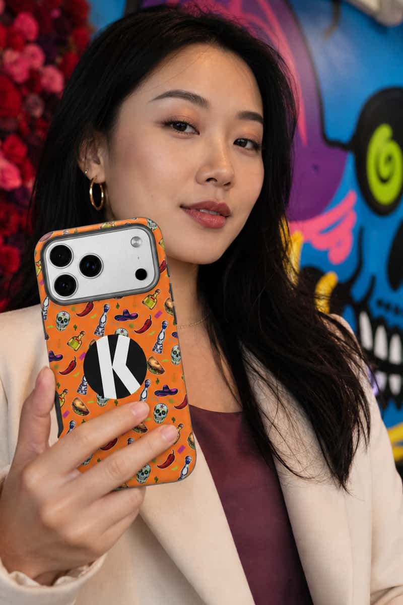 A woman holding a smartphone with an orange patterned phone case featuring a black circular logo with a white letter 'K'. The background shows a colorful graffiti wall.