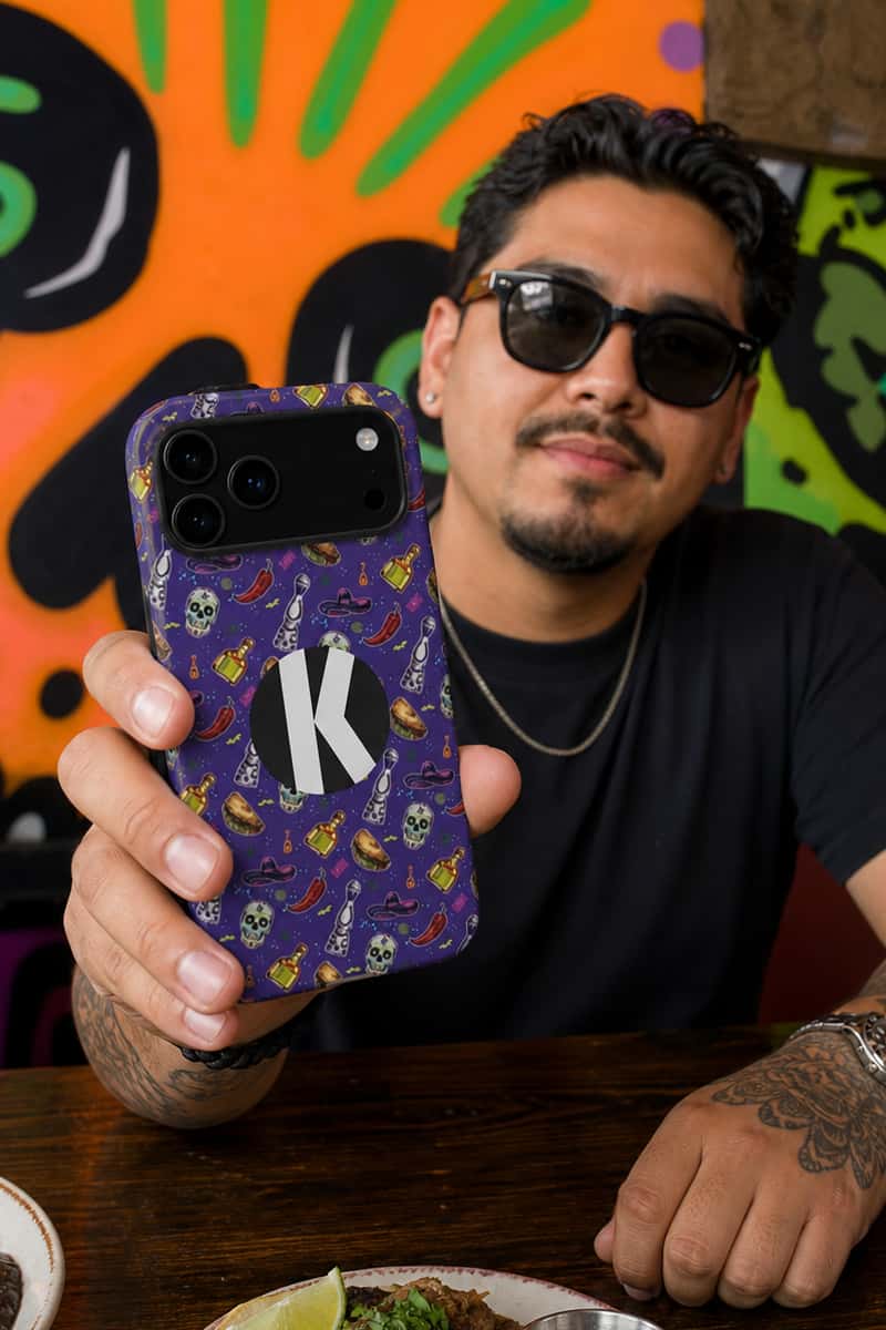 A man wearing sunglasses and a black t-shirt holds up a purple phone case decorated with a pattern of skulls, chili peppers, and a central white circle containing a black letter 'K'. The background features a vibrant, colorful graffiti wall.