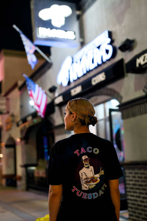 Woman wearing black Kalaveras Taco Tuesday Tee back view outside a Kalaveras restaurant at night